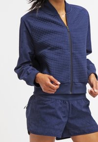 Chaqueta bomber azul marino con textura acolchada y un acabado suave, cierre de cremallera, puños acanalados, combinada con pantalones cortos a juego.