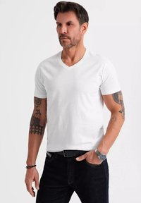 T-shirt bianca a scollo a V realizzata in cotone, maniche corte, con un design aderente. Il modello ha dei tatuaggi e indossa jeans scuri e un orologio d'argento.