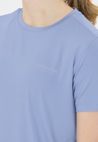 Ljusblå sportskjorta med kort ärm och texten "ENDURANCE" på bröstet, bärs av en person med ljus hy och blont hår i hästsvans.