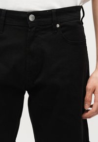 Κοντινό πλάνο ενός ατόμου που φορά μαύρο παντελόνι με ασημένιο κουμπί και νίκελ, που στέκεται μπροστά σε λευκό φόντο.