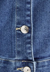 Μπλε τζιν ύφασμα με ορατές ραφές, δύο μεταλλικά κουμπιά και μια μικρή φθορά κοντά στο κουμπί.