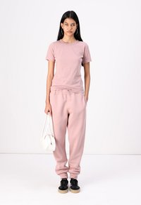 Camiseta de algodón rosa claro combinada con pantalones de chándal a juego. La modelo sostiene una bolsa blanca y lleva zapatillas negras. Diseño simple y casual.
