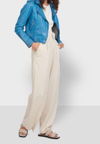 Chaqueta de cuero azul estilo biker con cremalleras plateadas, combinada con pantalones de tiro alto en color crema y sandalias trenzadas en tonos neutros.