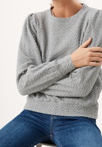 Grauer, strukturierten Pullover mit Fischgrätenmuster, rundem Ausschnitt und gerippten Bündchen. Kombiniert mit blauen Jeans, die Arme vor der Brust verschränkt.