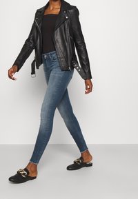 Chaqueta de cuero negra de motorista, top negro ajustado, jeans skiny azul medio, zapatos negros sin cordones con detalle de cadena dorada, caminando sobre un fondo liso.