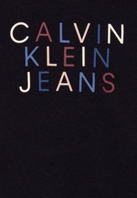 Nome del marchio Calvin Klein Jeans con lettere bianche, blu e bordeaux su sfondo di tessuto nero.