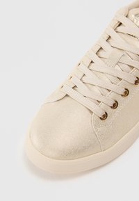 Nærbilde av beige sneaker tå med stofflisser som går gjennom gulløyler mot en enkel hvit bakgrunn.