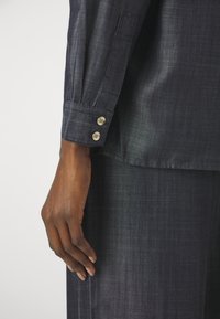 Camicia in denim scuro con maniche lunghe, presenta un polsino con bottoni e una trama liscia. Due bottoni visibili sul polsino sono di colore beige opaco.