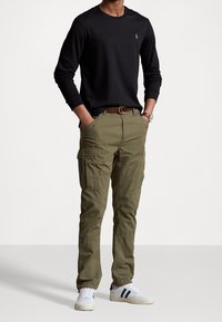 Man draagt een zwart shirt met lange mouwen, olijfgroene cargobroek met meerdere zakken, witte sneakers met donkere strepen en een bruine riem.