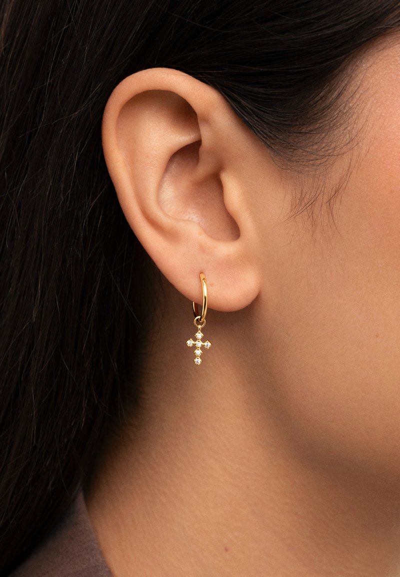 Boucle d'oreille en or avec un design de croix ornée de petites pierres décoratives, suspendue à un fin anneau, présentée sur un teint de peau.