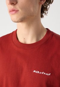 Raudona medvilninė džemperis su įsiuvamu apykaklės kraštu ir baltu siuvinėtu "MARCO POLO" logotipu krūtinėje. Lygus paviršius be raštų.