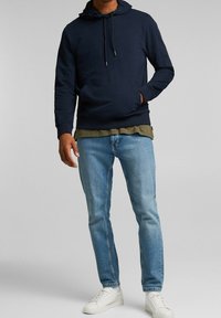 Sudadera con capucha de color azul marino con cordones, combinada con una camiseta de color verde oliva desvanecido y jeans de color azul claro. Zapatillas blancas completan el conjunto.