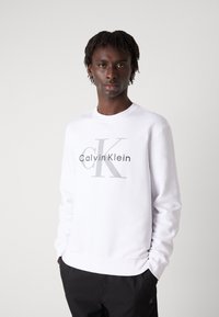 Felpa bianca realizzata in misto cotone, con un grande logo grigio di Calvin Klein centrato sulla parte frontale, polsi e orlo a coste.