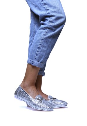 Scarpe intrecciate argento con tacco piatto e hardware decorativo, abbinate a jeans azzurri risvoltati. Sfondo bianco.