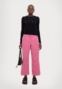 Maglione nero a maniche lunghe con ornamenti; pantaloni a gamba larga in velluto rosa; persona che tiene una piccola borsa nera; sandali neri chunky.