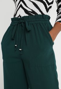 Pantalons verts avec ceinture élastique et cordon de serrage, dotés de pointes métalliques. Texture lisse avec poches latérales. Pas de motifs ni d'accents visibles.