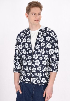 Hanorac cu fermoar navy cu model floral de hibiscus alb, țesătură ușoară, mâneci lungi și buzunare frontale, purtat peste o cămașă albă.