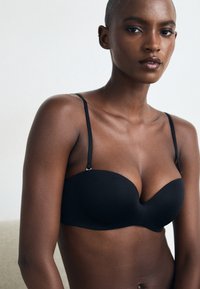 Soutien-gorge noir sans bretelles avec des bonnets lisses et galbés, des bretelles fines ajustables et un design sans coutures. Fini mat et détails matériels minimaux.