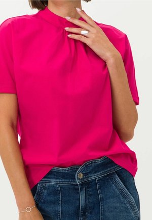 Woman wearing bright pink, short-sleeve blouse with high neckline and dark blue denim jeans, hand near collar showing silver ring and bracelet.