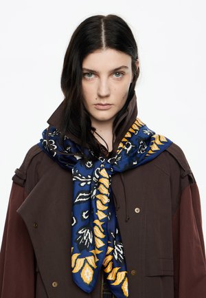 Bufanda de seda azul con patrones florales amarillos y negros, atada de forma ordenada alrededor del cuello, combinada con una chaqueta exterior marrón.