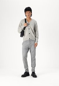 Grauer Strickcardigan mit V-Ausschnitt und schwarzen Knöpfen, kombiniert mit grauen karierte Hosen und schwarzen Schnürschuhen. Lässige Umhängetasche.