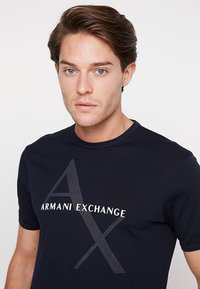 Marineblå bomulls t-skjorte med trykket hvitt "AR MANI EXCHANGE"-logo og et stilisert "A"-design. Korte ermer, rund hals.