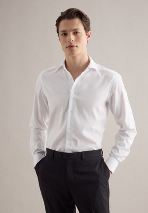 Junger Mann steht mit den Händen in den Taschen, trägt ein weißes Hemd mit Knöpfen und schwarze Hose vor einem einfarbigen Hintergrund.