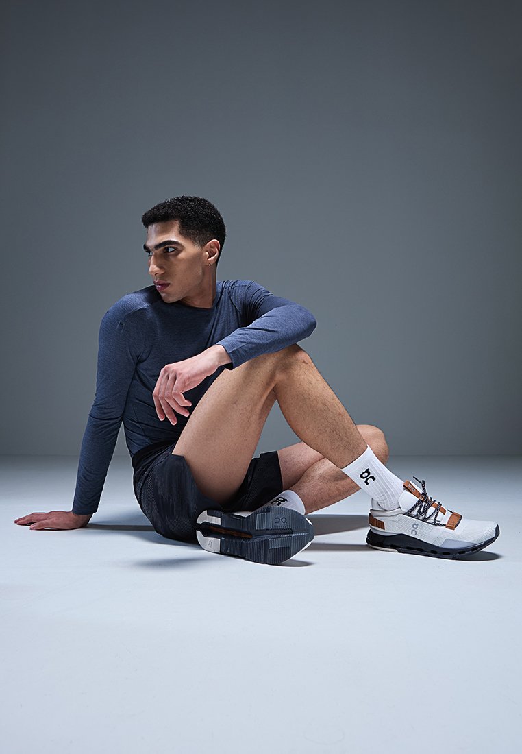 Sportliches Schuhwerk mit einem weißen und grauen Obermaterial, braunen Akzenten und einer strukturierten Sohle. Modell in einem langärmeligen navyblauen Oberteil und schwarzen Shorts, sitzend.