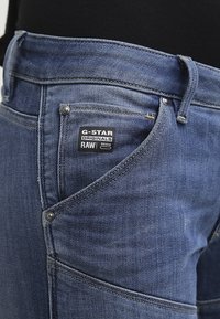 Jeans de mezclilla azul con una ligera textura desgastada, que presentan una etiqueta de G-Star Originals, accesorios metálicos y detalles de costura prominentes.