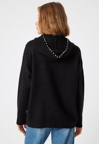 Schwarzer Strick-Hoodie mit lockerer Passform, der einen Perlenakzent entlang des Randes der Kapuze aufweist. Die Textur ist weich und wird durch Rippbündchen und Saum ergänzt.