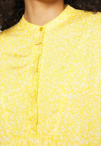 Gul blomstret bluse med rund halsudskæring, knaplukning og let rynkning ved kanten. Letvægts bomuldsstof.