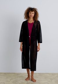 Must mustriga kimonotüüpi katteese musta võrgu riietus, millel on pikad varrukad ja krae, ning vöödetail taljes. Kandmine tumesinise ujumistrikoo üle.