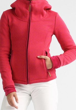 Vrouw draagt een rode, getextureerde jas met rits en zijzakken en witte broek, hand in één zak.