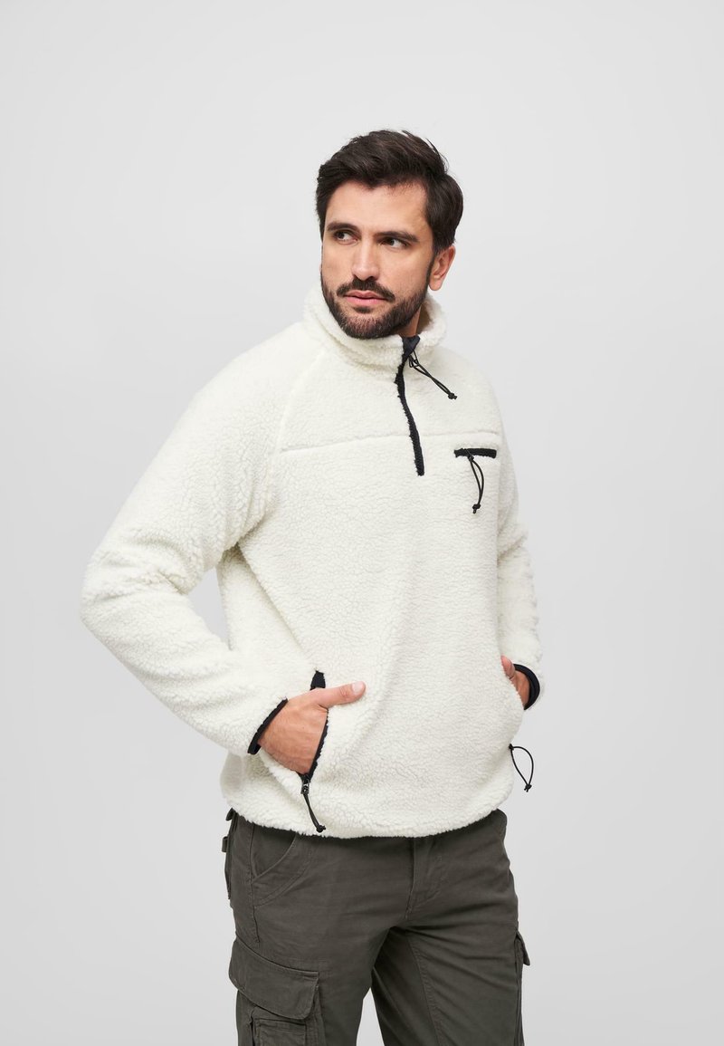 Polarowy golf w odcieniu off-white z wysokim kołnierzem, czarną zapinaną na zamek kieszenią na klatce piersiowej oraz regulowanym dołem; ma czarne akcenty i ściągacze na rękawach.
