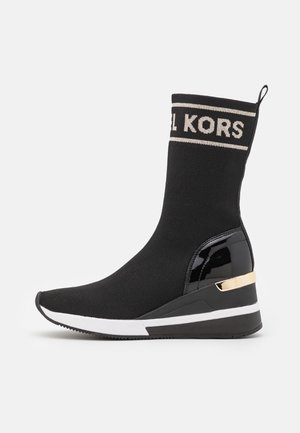 Czarny but typu sneaker w stylu skarpety sięgający do połowy łydki, z białą podeszwą, złotym detalem na obcasie, lakierowaną panelem na pięcie oraz taśmą z logo „KORS” na górze.