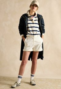 Jūras flotes vieglais žakets, balta krekla ar jūras flotes svītrām, baltas šortus, zilo un balto sporta apavu, un bēša cepure. Stāvošā pozā.