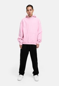 Rosa Kapuzenpullover mit einer Fronttasche, lockerer Passform und Kordelzugkapuze, kombiniert mit schwarzen Sweatpants und weißen Sneakers. Einfaches, ungeschmücktes Design.