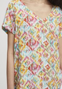 Blouse multicolore avec un col en V, présentant un motif abstrait texturé en forme de diamant dans des nuances de rouge, jaune, bleu et blanc, à manches courtes.