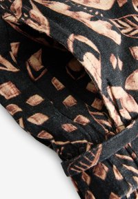 Tissu noir avec des motifs géométriques et de feuilles marron, montrant un élastique froncé et des coutures cousues en gros plan.