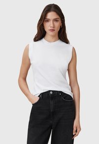 Weißes ärmelloses Oberteil aus Baumwolle mit rundem Ausschnitt, kombiniert mit hochgeknöpften schwarzen Jeans im klassischen Fünf-Taschen-Design.