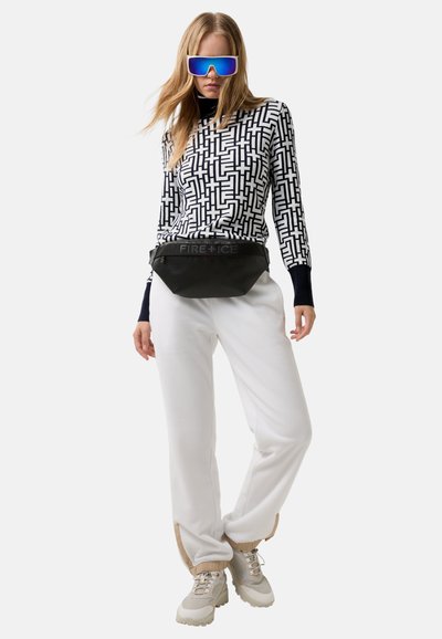 Kobieta ubrana w geometryczną czarno-białą koszulkę, białe spodnie, masywne sneakersy, lustrzane niebieskie okulary przeciwsłoneczne oraz czarną torbę na pas z napisem "FIRE + ICE".