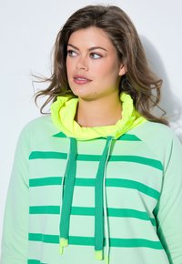 Mujer con cabello castaño ondulado, vistiendo una sudadera con capucha de rayas verdes y verde claro, con un cuello fruncido de neón amarillo, mirando ligeramente hacia la izquierda.