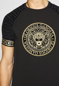 Camiseta negra con emblema de león dorado y texto "GLORIOUS GANGSTA". Presenta mangas de patrón negro texturizado y un cuello redondo.