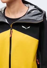 Άτομο με κίτρινο και μαύρο μπουφάν με κουκούλα, γκρι επένδυση και εμπρόσθιο φερμουάρ με πορτοκαλί κορδόνια.