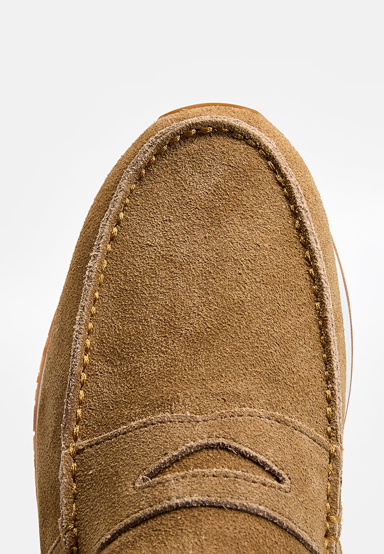 Chaussure en daim marron avec un bout arrondi, présentant des coutures visibles le long des bords et une surface lisse et texturée. Construction légère.