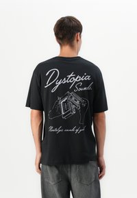 Mann iført svart t-skjorte med hvit grafikk av hender som holder kassettbånd og teksten "Dystopia Sounds. Nostalgiske lyder av ungdom."
