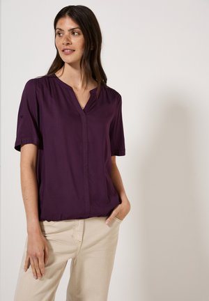 Blouse violette à manches courtes avec un décolleté en V, présentant des fronces discrètes aux épaules et une coupe décontractée, associée à un pantalon beige clair.