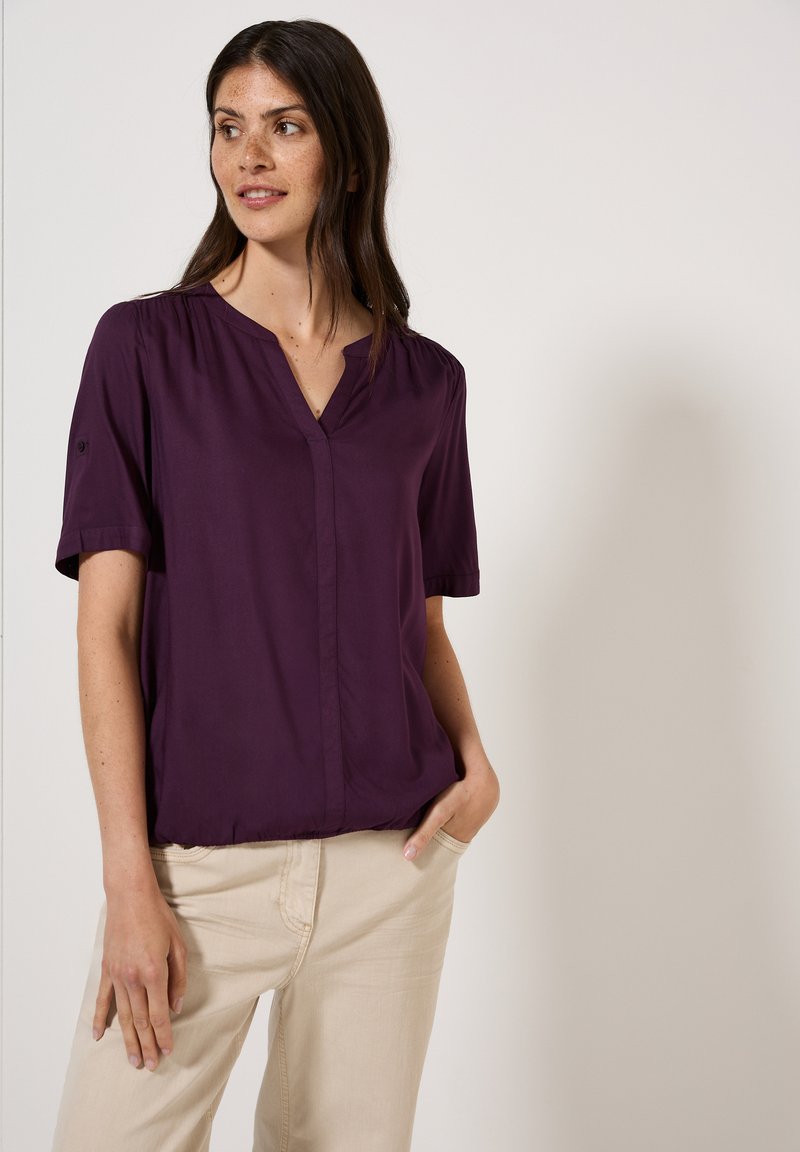 Blusa corta viola a maniche corte con scollo a v, caratterizzata da una leggera arricciatura sulle spalle e una vestibilità morbida, abbinata a pantaloni beige chiari.