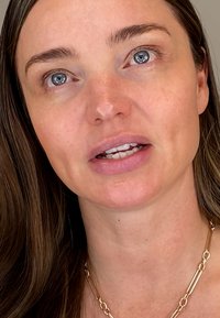 Close-up of a face with medium-length brown hair, wearing minimal makeup. A gold chain necklace is visible on the neck.