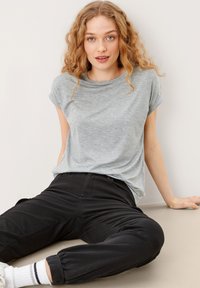 Jasnoszara, krótki rękaw t-shirt z okrągłym dekoltem, w połączeniu z czarnymi spodniami. Tkanina jest miękka, a krój luźny. Siedzący na neutralnej powierzchni.